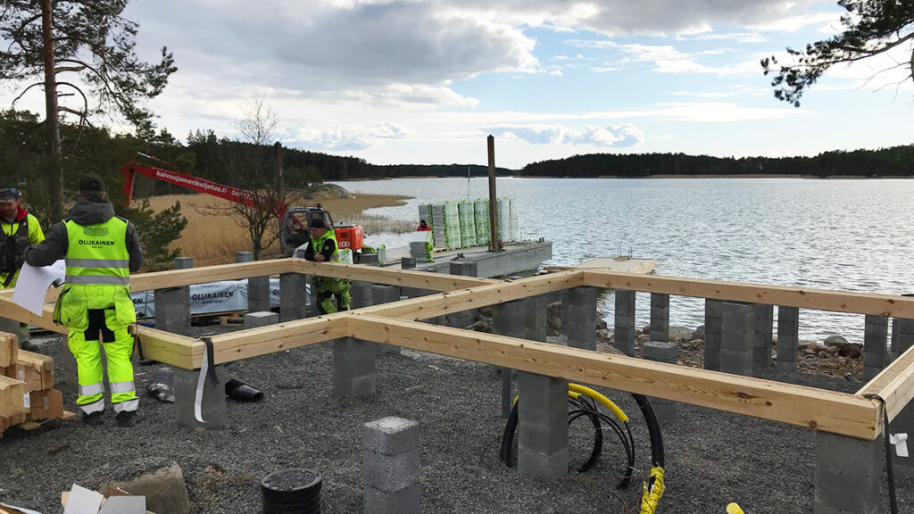 Kuvassa Ollikaisen hirsirakenteen rakentajat pystyttämässä hirsitaloa saaristossa.